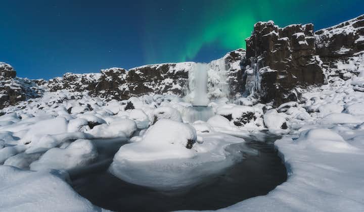 The Oxararfoss waterfall at Thingvellir National Park looks breathtaking surrounded by a snow-covered landscape with the northern lights overhead.