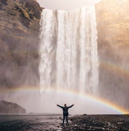 A person stands in front of the mighty Skogafoss waterfall on Iceland's South Coast with their arms stretched wide as a rainbow graces the front of the cascade.