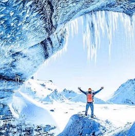 A person stands in the entrance of an ice cave in Iceland with their arms stretched out and spectacular scenery surrounding them.