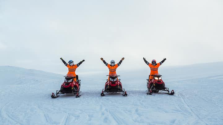 People driving snowmobiles across the Langjokull glacier stop for a photo with their arms in the air amid a vast icy expanse.