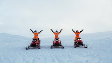People driving snowmobiles across the Langjokull glacier stop for a photo with their arms in the air amid a vast icy expanse.