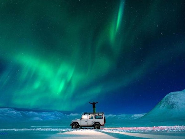 A person stands on a jeep amid a snow-covered landscape with the northern lights above, close to Soti Lodge on the Trollaskagi P