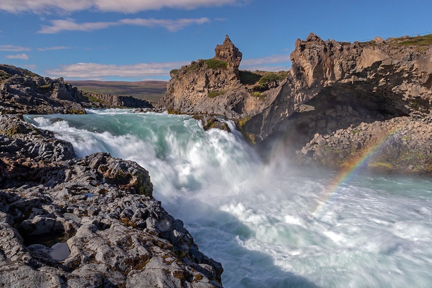The Geitafoss waterfall in North Iceland, seen on a sunny day with a rainbow above it.