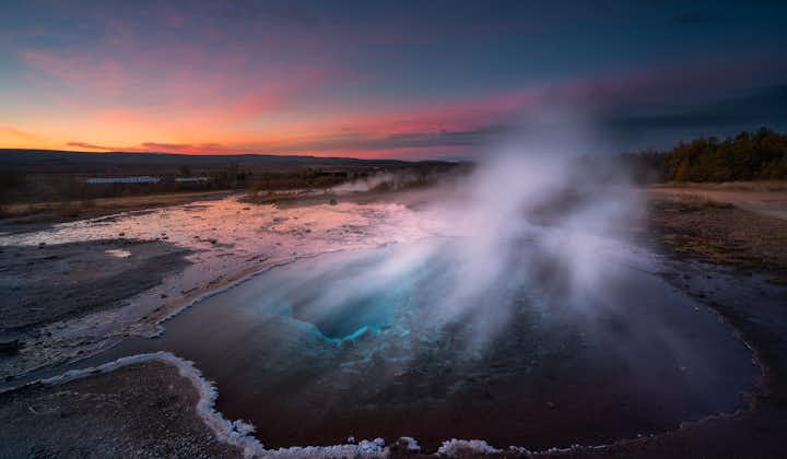 The Geysir geothermal area looks surreal at sunset with its misty atmosphere and vibrant colors swirling around.