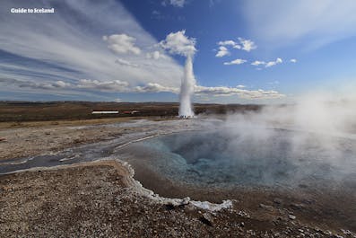 The Strokkur geyser erupts high into the air at the Geysir geothermal area on a cloudy, blue-sky day.
