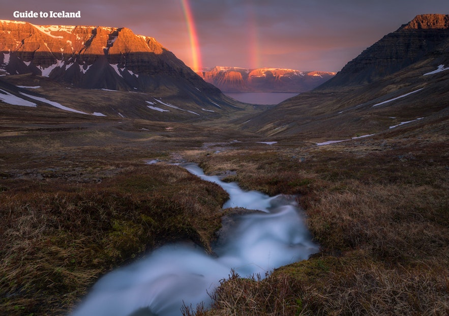 In the Westfjords of Iceland, a silken river rushes through a snow-capped mountains under a rainbow at night.