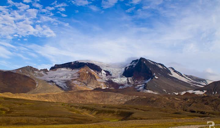 A view of jagged, snow-capped mountains, rolling green-brown terrain, and a glimpse of a road in East Iceland on a blue-sky day.