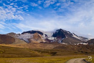 울퉁불퉁한 눈 덮인 산과 구불구불한 녹갈색 지형, 그리고 푸른 하늘이 펼쳐진 날 아이슬란드 동부의 도로를 엿볼 수 있는 풍경.