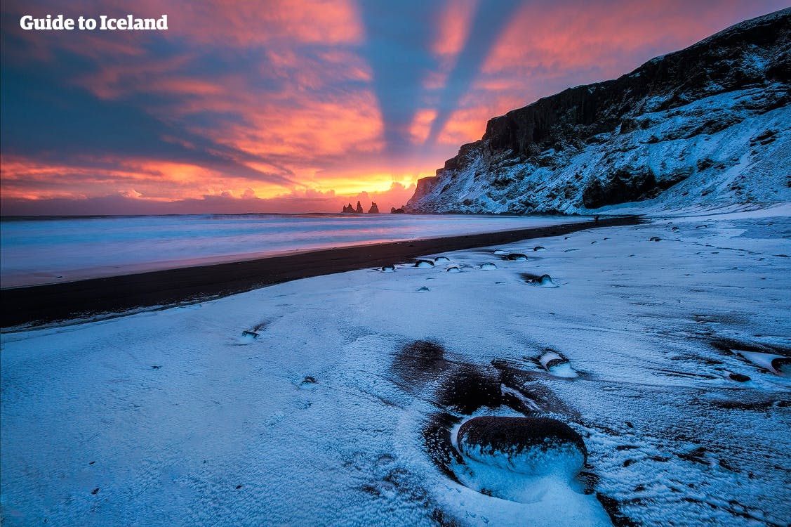 Reynisfjaras svarta sandstrand ligger täckt i snö medan vintersolens sista strålar målar himlen röd.