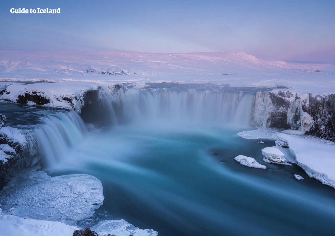 Im rosafarbenen Licht der tiefstehenden Wintersonne kämpft der Goðafoss-Wasserfall gegen die eisigen Elemente und fließt weiter durch die verschneite Landschaft im Norden Islands.
