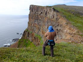 A hiker stands on grassy terrain on top of the green cliffs of Hornstrandir inthe Westfjords.