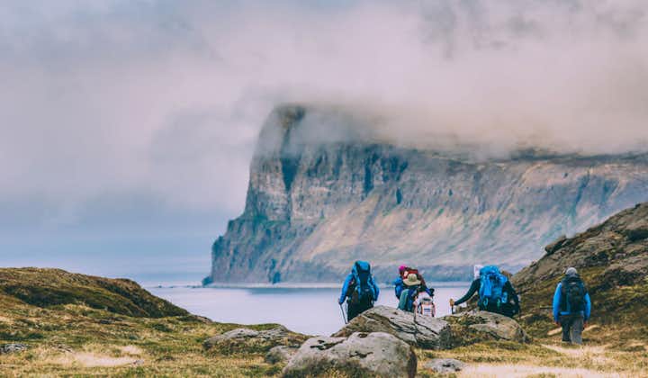 Hikers explore the beautiful Hornstrandir Nature Reserve with calm fjord waters in front and sheer cliffs rising behind, partially enveloped by cloud cover.