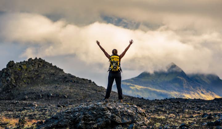A person standing on the Thrihyrningur mountain summit stands with their arms raised in the air on a cloudy day.