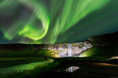The lorthern lights moving like dancers in the sky above a waterfall.