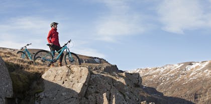 En kvinde står ved siden af en mountainbike på toppen af en klippe iført hjelm og regntæt rød jakke.