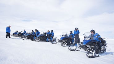Four people, three of them on snowmobiles, wearing safety overalls and helmets.