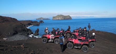 Group ATV tour stopping on a black sand beach with the ocean and rocky outcrops in front.