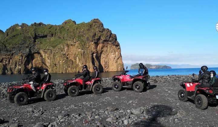 On a blue-sky day, ATV riders on a black sand beach in Vestmannaeyjar with a large rocky outcrop behind.