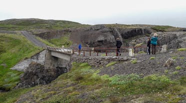 Folk går over en bro midt i et barskt fjordlandskab på Østisland.