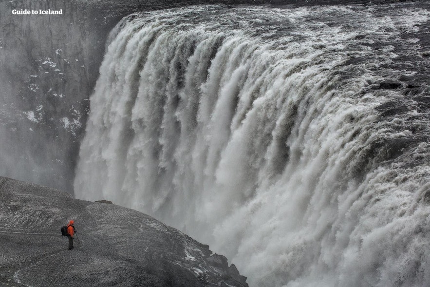 Ein Mann, der auf einer Klippe steht, sieht im Vergleich zum nahe gelegenen Dettifoss-Wasserfall winzig aus.