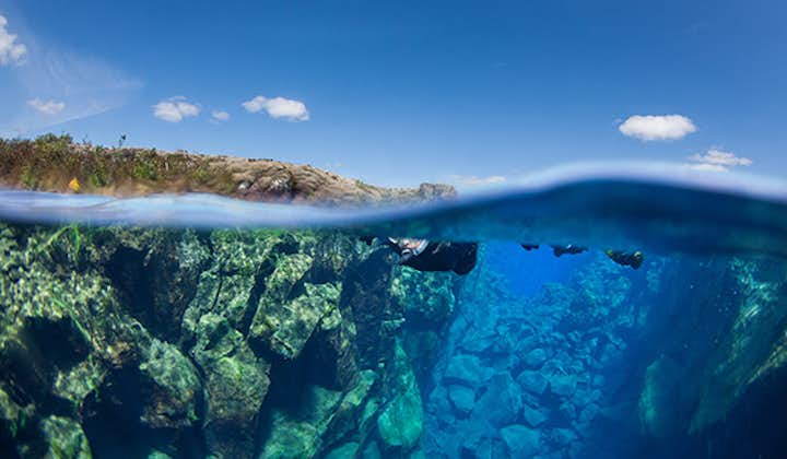 People are snorkeling through the crystal-clear water at the Silfra fissure amid an underwater landscape of blue and green colors.