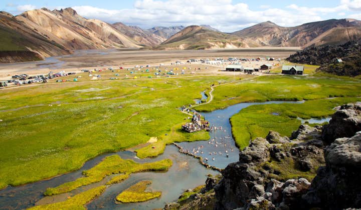 A birdseye view of Landmannalaugar mountain with people bathing in the natural hot spring, tents dotted around the plains, and the mountains towering around.