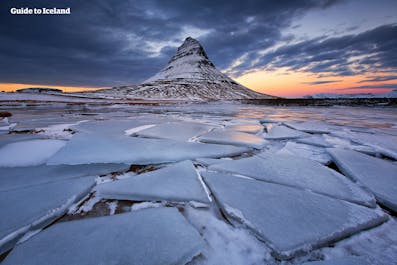 Mount Kirkjufell on the Snaefellsnes Peninsula in the West of Iceland. In the foreground, the surface of a lake is covered with cracked pieces of ice.