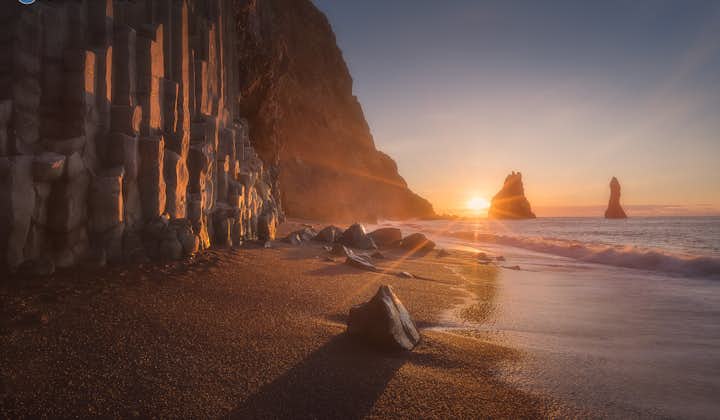 The sun peaking over the horizon over Reynifjara Black Sand Beach. Hexagonal basalt rock columns can be seen in the foreground to the left.