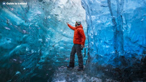 En reisende i knallrød frakk står i en blå og hvit isgrotte på Island.