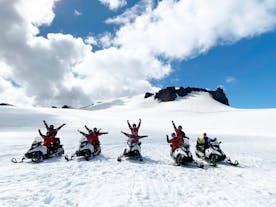 A group of people are seen sitting on snowmobiles on top of Vantajokull Glacier with their hands up in the air.