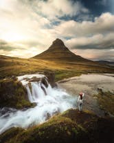 Woman wearing bobble hat standing looking at Kirkjufell mountain and waterfall.