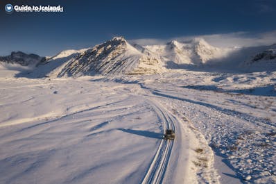 A vehicle traverses the vast, snow-covered landscapes of East Iceland on a bright day with the mountains in the background.