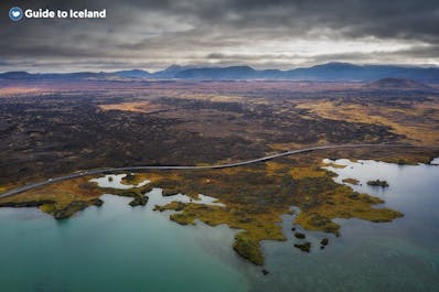 A birds eye view of the edge of Lake Myvatn with its calm water and a striking, colorful landscape stretched out in front.
