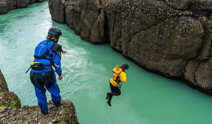 A person prepares to jump from the top of a rock on the Hvita river as they watch the person in front in mid-air.