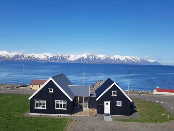Exterior view of Grenivik Guesthouse on a blue-sky day with the grass surrounding, sea behind, and mountains beyond.