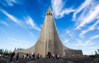 People walking in front of the tall tower of Hallgrimskirkja church on a cloudy blue-sky day.