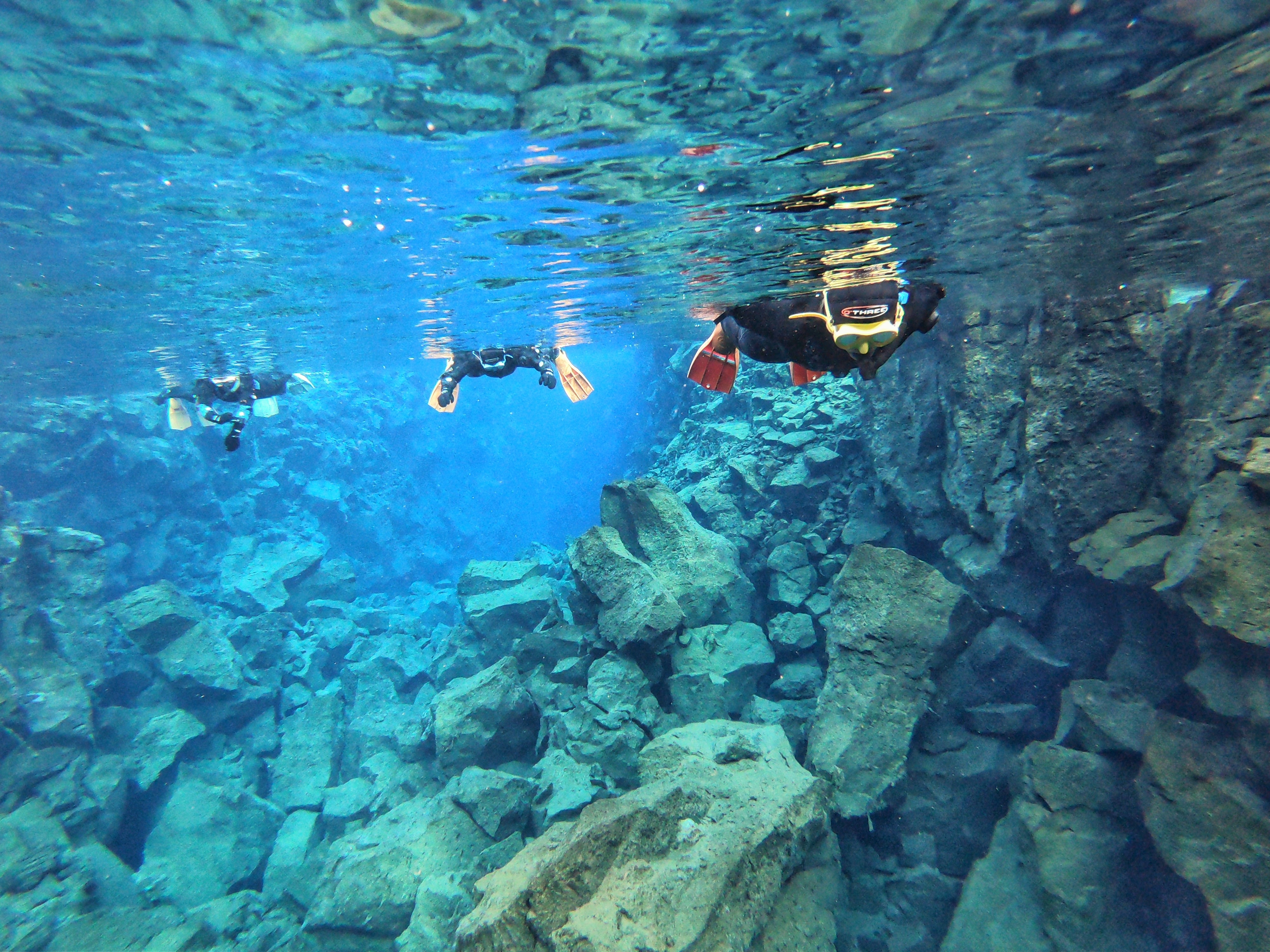 People snorkeling in the Silfra Fissure amid bright blue water and a rocky underwater landscape.