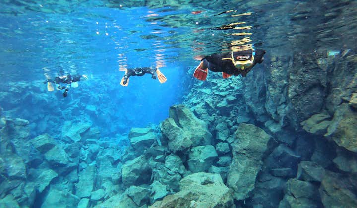 People snorkeling in the Silfra Fissure amid bright blue water and a rocky underwater landscape.