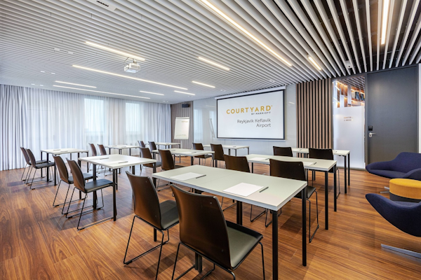 A meeting room with tables, chairs, a projector screen, and a presentation board at the Courtyard by Marriott Reykjavik Keflavik