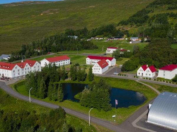A birdseye view of Hotel Laugar Reykjadalur, surrounded by green hills, with trees and a small lake in front.