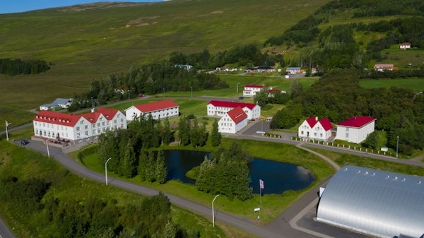 A birdseye view of Hotel Laugar Reykjadalur, surrounded by green hills, with trees and a small lake in front.