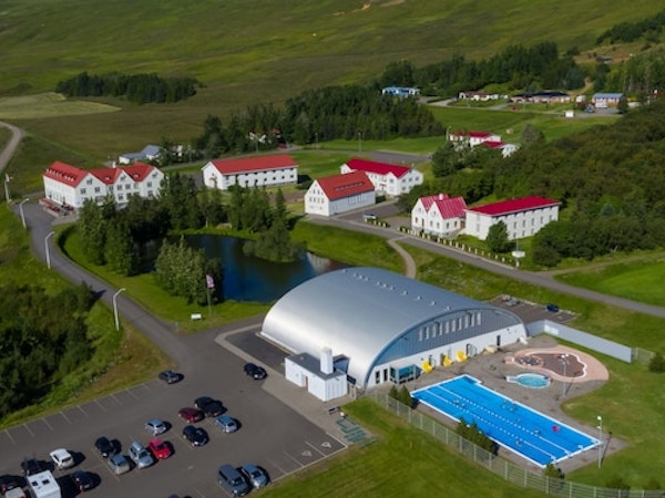 A birdseye view of Hotel Laugar Reykjadalur, surrounded by green hills, with trees, a small lake, and an outdoor geothermal pool