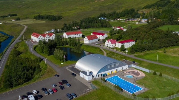 A birdseye view of Hotel Laugar Reykjadalur, surrounded by green hills, with trees, a small lake, and an outdoor geothermal pool
