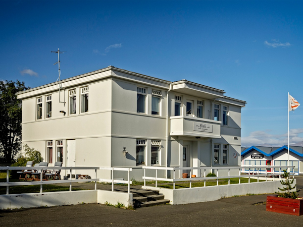 White exterior view of Dalvik HI Hostel with grass in front and a fence around on a sunny, blue-sky day.