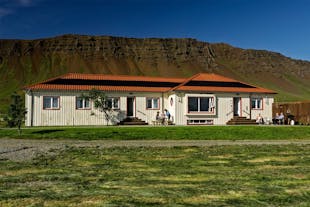 The exterior view of Reykholar HI Hostel with a grass area in front, the mountains directly behind, and people sitting around a 