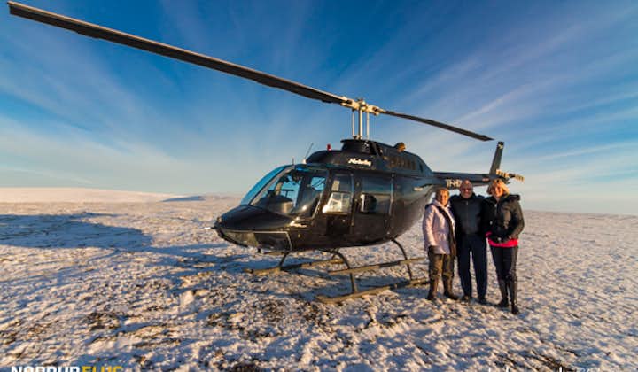 Three happy people are standing on the snowy terrain beside a black Nordurflug helicopter on a mountain landing near Reykjavik.