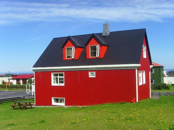 An exterior view of the red house at Grundarfjordur HI Hostel with a picnic table on the grassy surrounding area on a bright, bl