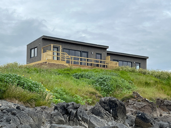 Exterior view of Eyri Seaside Houses in Northwest Iceland perched on a coastal hillside with grass and rocks below.