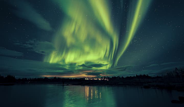 The aurora borealis lights up the starry winter night sky and town lights reflect onto the water below.