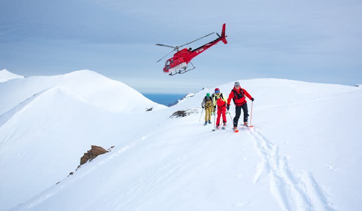 Three people skiing with a helicopter taking off behind them on a mountain in North Iceland.